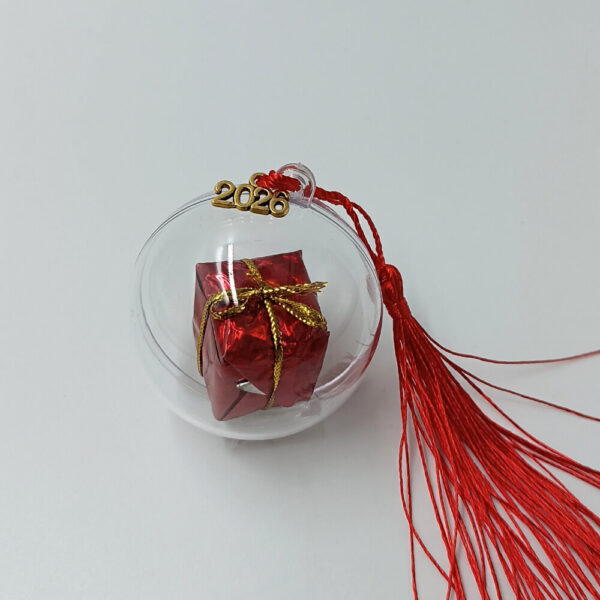 Διάφανο γούρι plexiglass με μικρό τυλιγμένο δώρο σε κόκκινο χαρτί και χρυσό κορδόνι, και στοιχείο 2026.